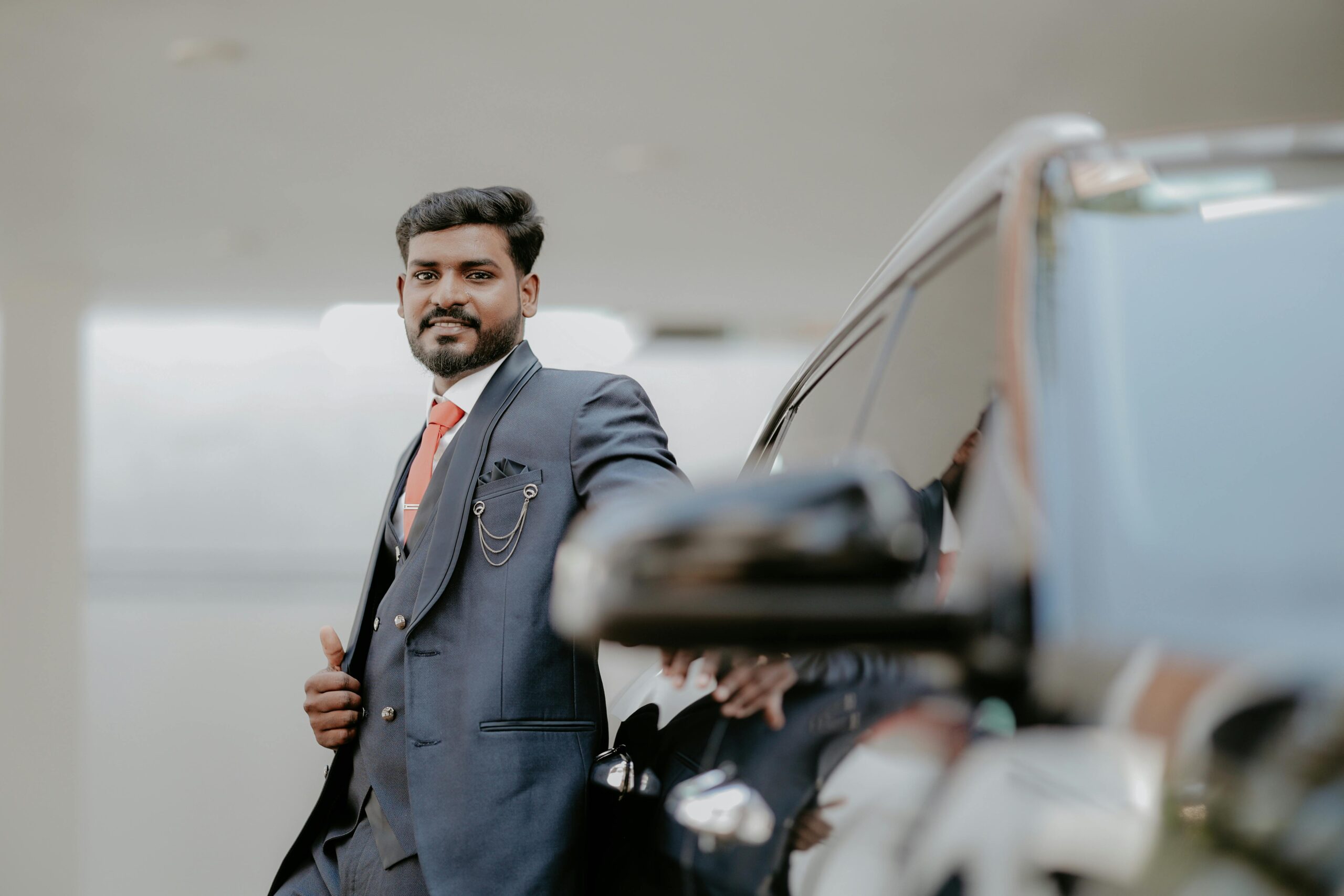 Confident Indian man in a suit posing beside a car, showcasing modern fashion and style.