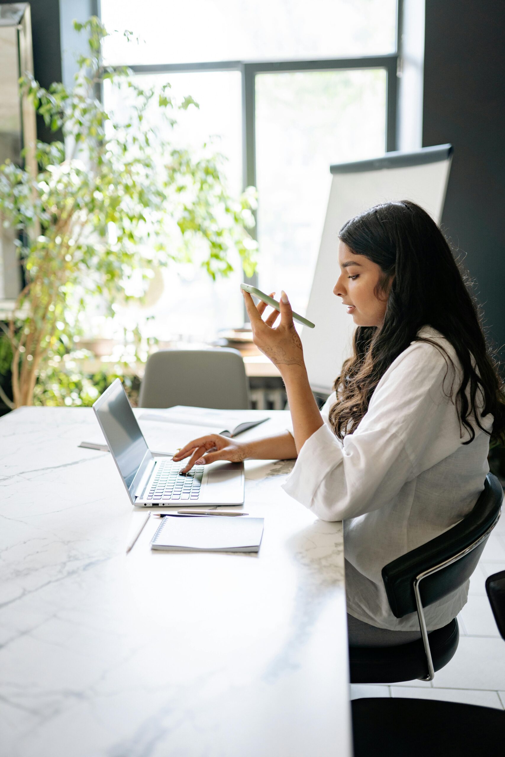 South Asian woman multitasking in a modern office using a laptop and mobile phone.